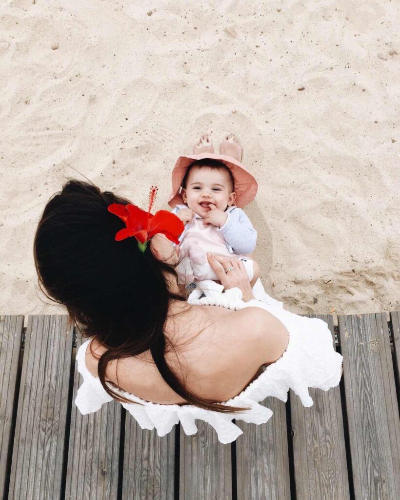 Image of a woman with a flower in her hair holding a baby in her lap on a boardwalk on a beach