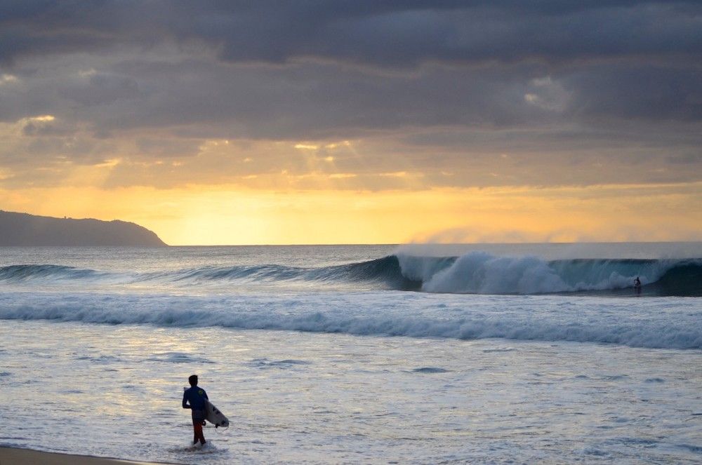 Banzai Pipeline in North Shore Oahu. Image of a man walking in the waves with a surfboard at sunset