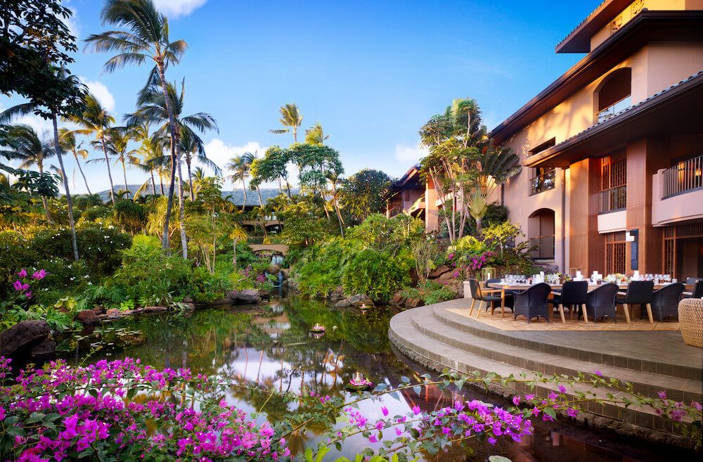 Image of a hotel with outdoor dining next to a stream with lots of green fauna