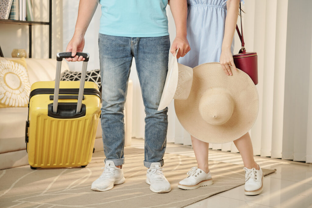 Get the best Hawaii packing list for couples created by top Hawaii blog Hawaii Travel with Kids. Image of oung couple with straw hats standing in room with suitcase ready for summer vacation