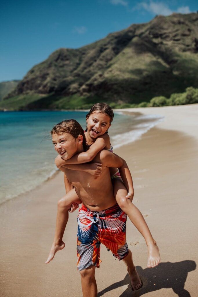 Image of a boy in a swimsuit giving a girl a piggyback ride on a beach in Hawaii