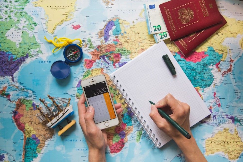 Image of someone using a calculator app on their phone with a map, notebook, compass, and passports