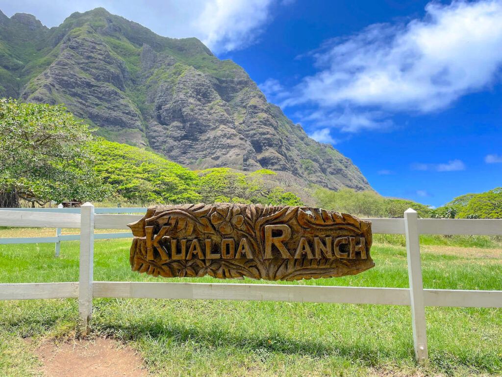 Image of a wooden Kualoa Ranch sign on a white fence with mountains in the background