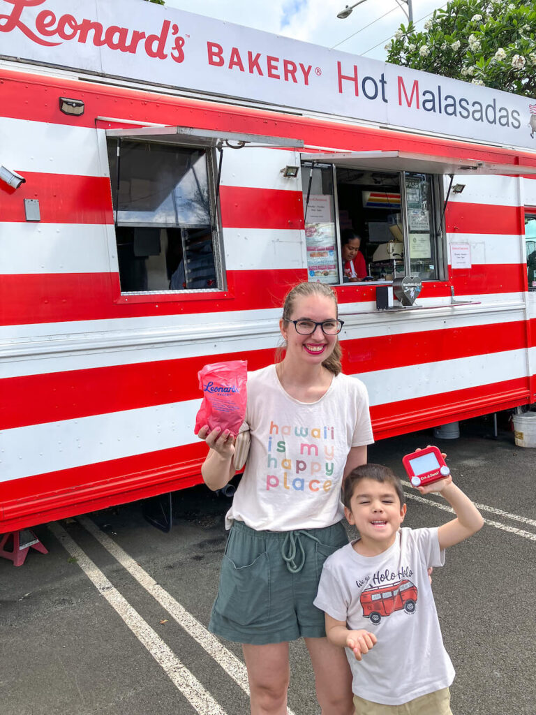 Image of a mom and son holding a bag of malasadas in front of a red and white striped Malasada food truck