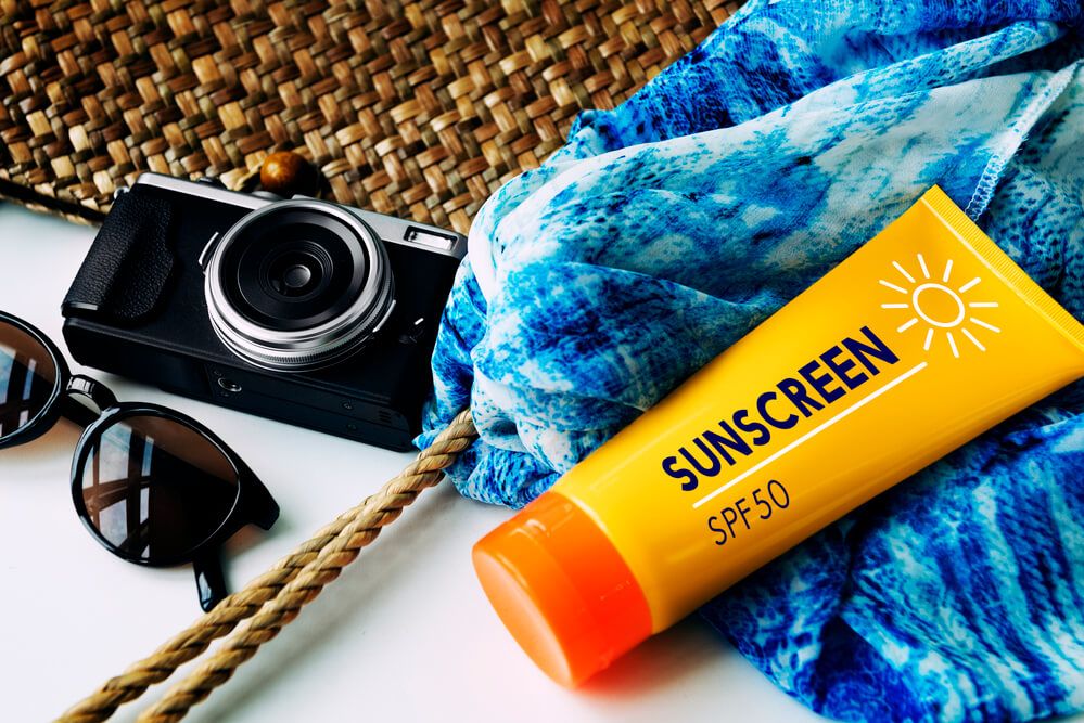 Image of sunscreen, a towel, a camera, sunglasses, and a straw bag