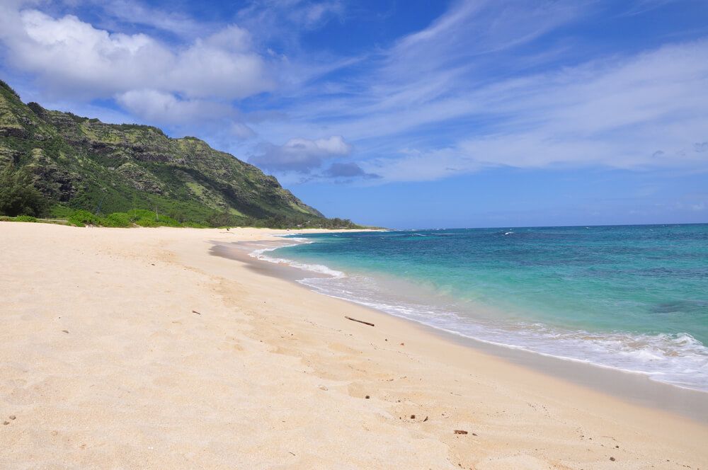 Image of a white sandy beach on Oahu with mountains in the background.