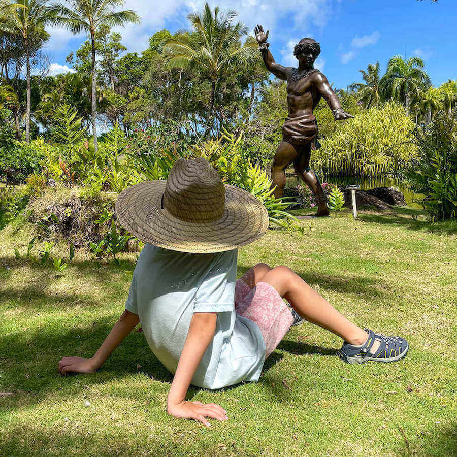Image of a boy sitting on the grass at Na Aina Kai Botanical Garden on Kauai with a hula statue in the background.