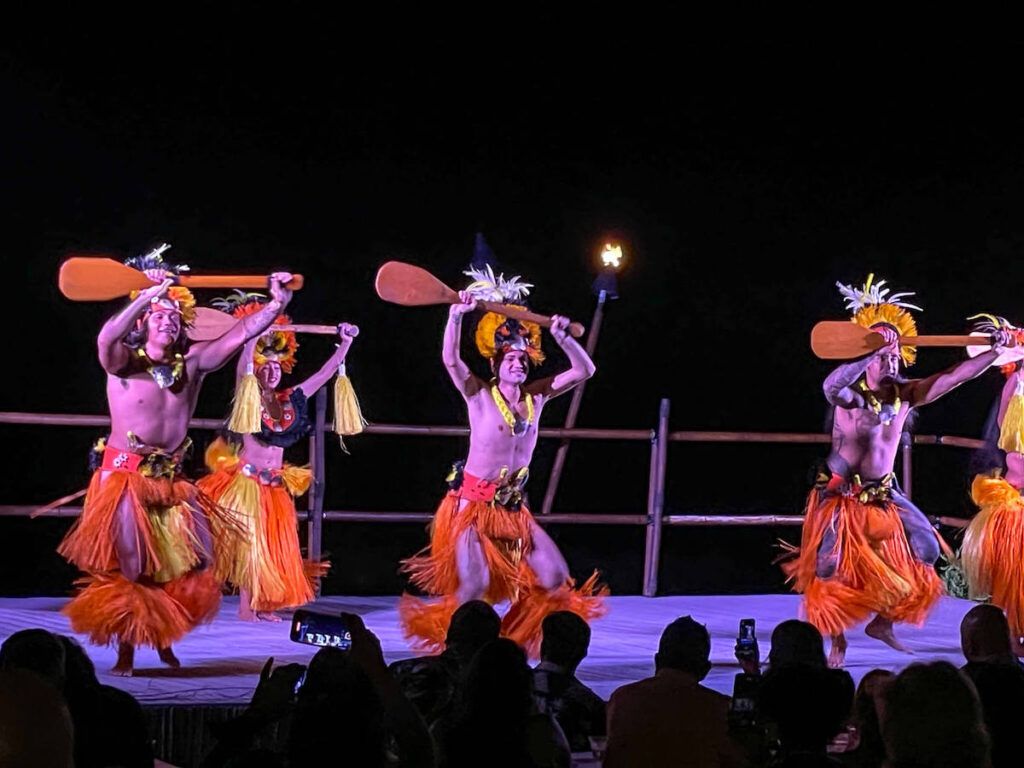 Image of men dancing with orange skirts and wooden paddles at the Voyagers of the Pacific Luau in Kona.