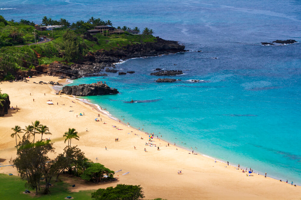 Waimea Bay Beach Park in North Shore Oahu. Image of a wide sandy beach with people on it and bright blue ocean water.