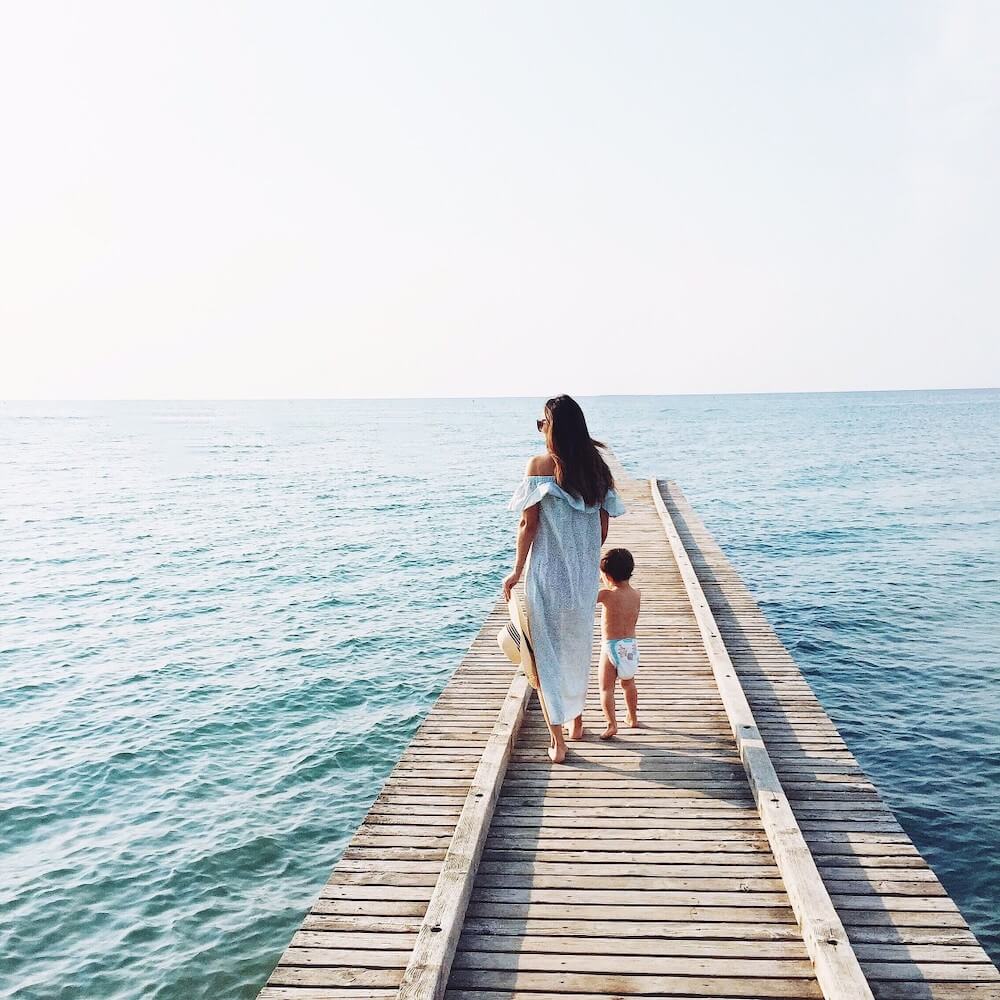 Image of a mom holding hands with a baby wearing a diaper as they walk on a boardwalk over the ocean