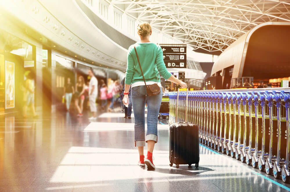 Image of a lady wearing jeans, red converse shoes, and a green sweater rolling a carry on bag through an airport