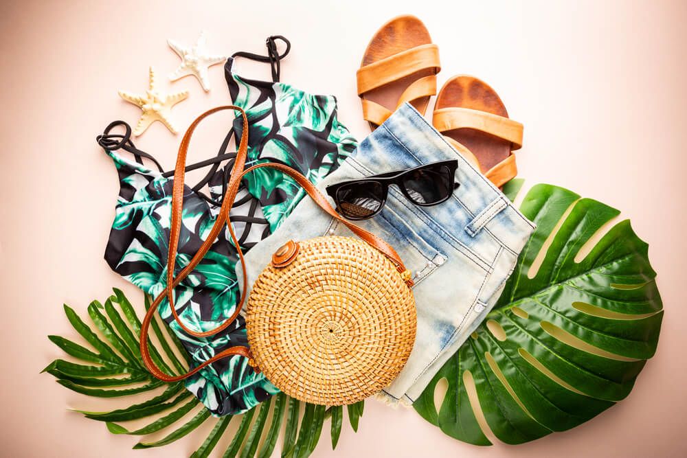 Image of monstera leaves with denim shorts, a tropical swimsuit, brown sandals, sunglasses, and a straw purse