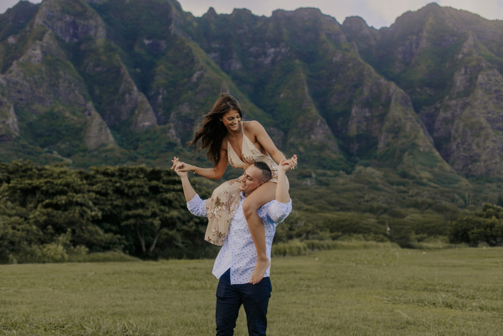 Image of a woman wearing a dress on the shoulders of a man in a button down shirt in front of Oahu mountains