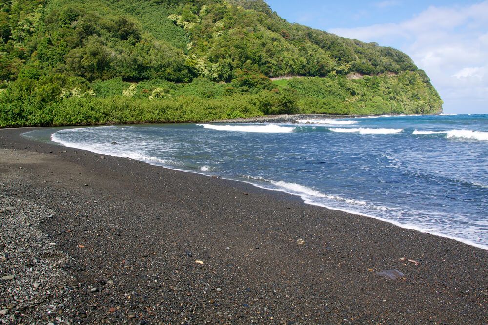 Hana Black Sand Beach at Waianapanapa State Park on Maui. Image of a black sand beach with a green hill in the background.