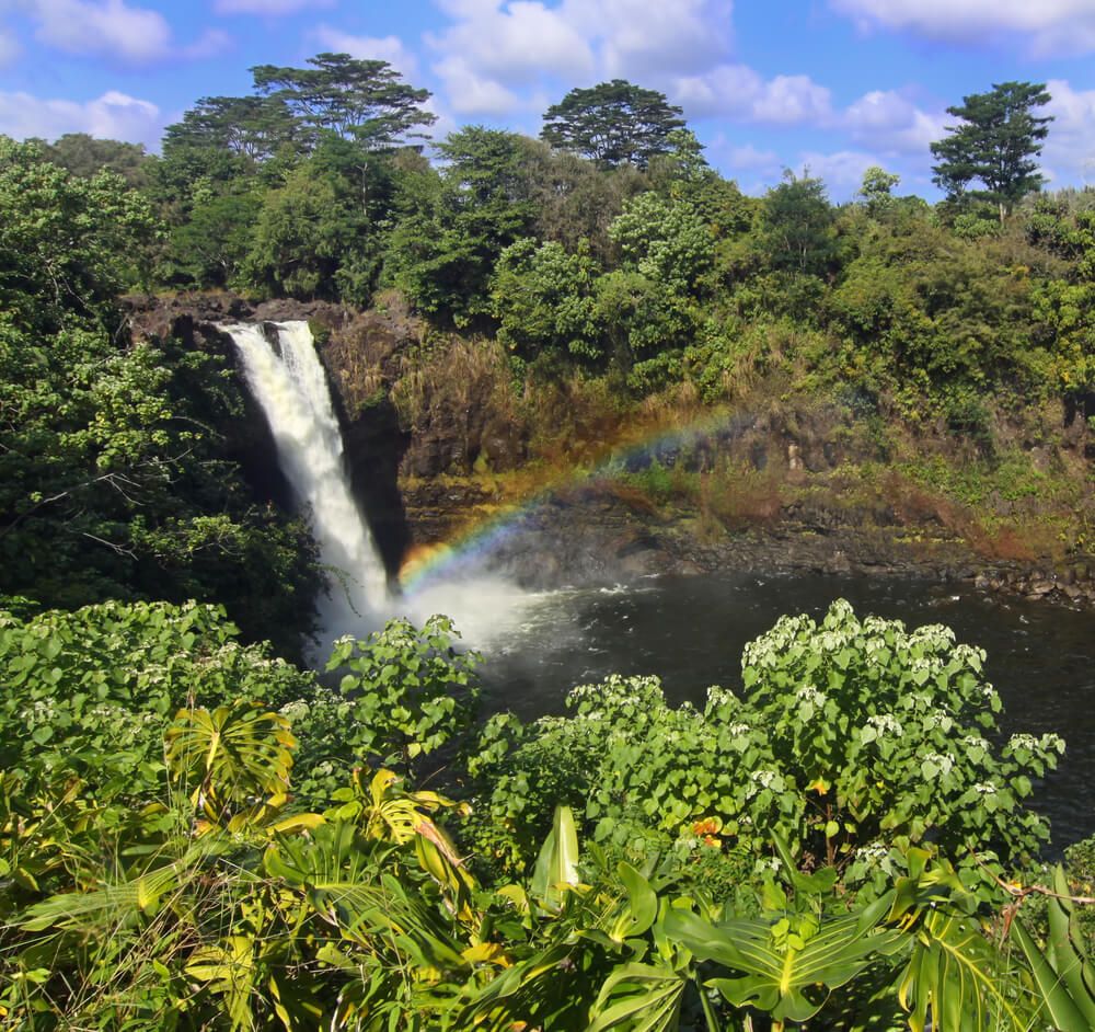 Rainbow Falls on the Big Island Hawaii. Image of a short waterfall going into a pool of water surrounded by greenery with a rainbow over the water