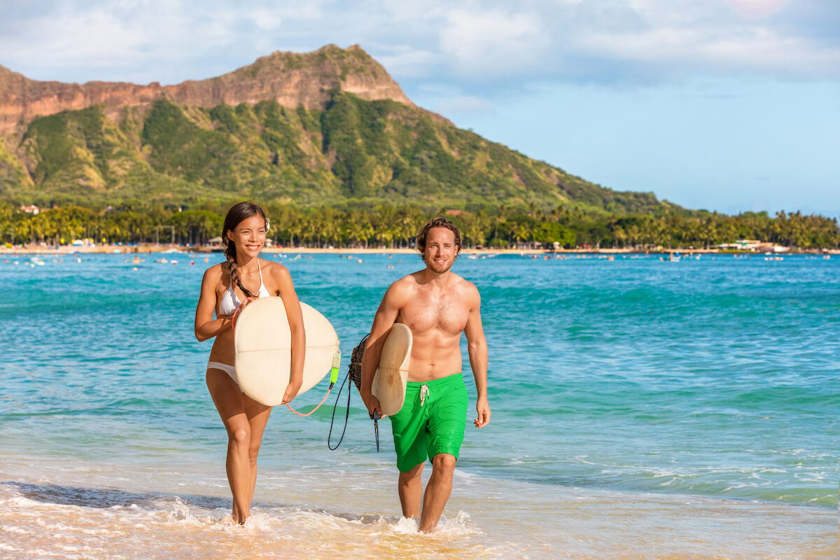 Find out the best things to do in Waikiki recommended by top Hawaii blog Hawaii Travel Spot! Image of a man and woman holding surfboards at Waikiki beach with Diamond Head in the background