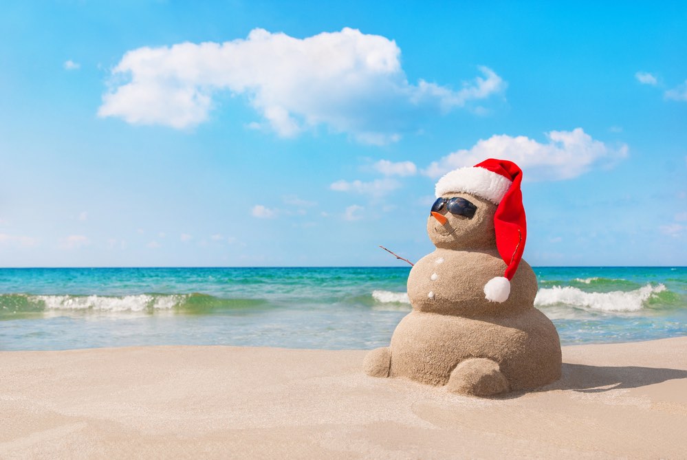 Image of Sandy christmas snowman in red santa hat and sunglasses at sunny beach. Holiday concept for New Years Cards.