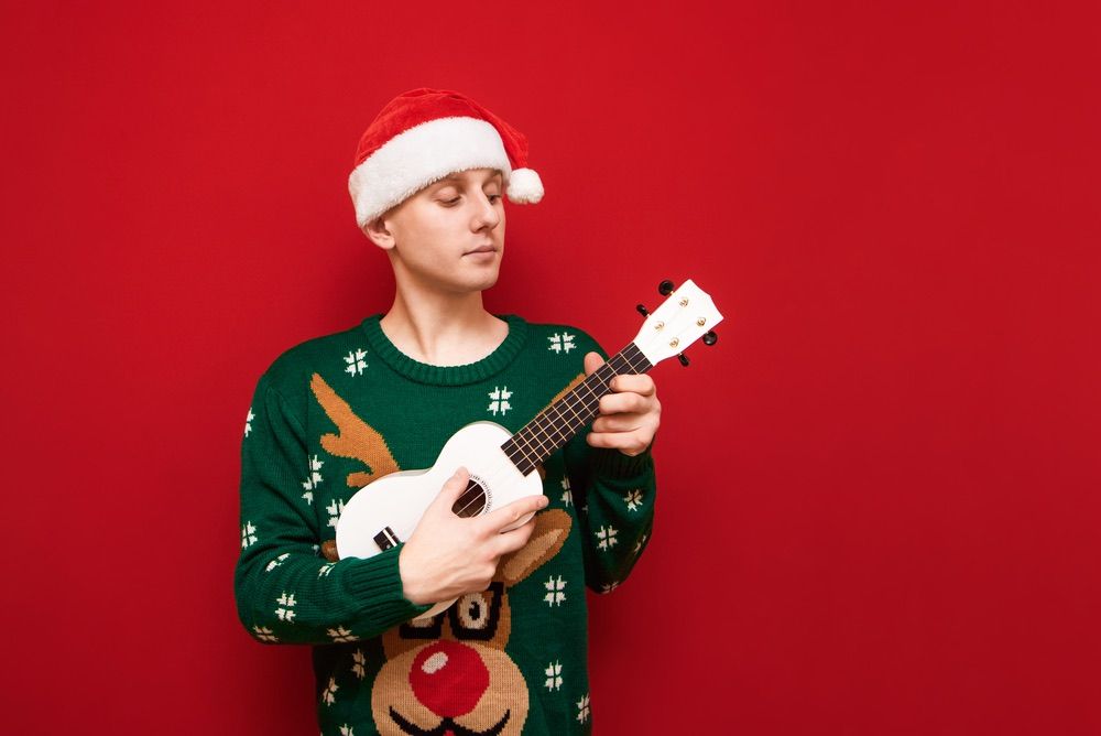 Image of Handsome young man in santa hat and warm sweater plays on ukulele on red background, looks away with serious face. Musician plays a Christmas tune not a ukulele. Isolated.