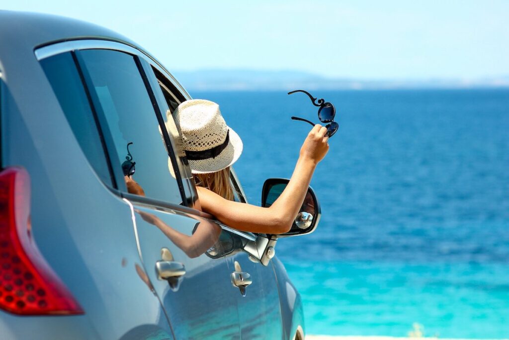 Renting a car is one of the easiest Kona to Hilo drive options. Image of a woman wearing a hat and holding sunglasses out a car window with the ocean in the background