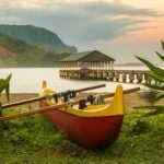 Check out these amazing things to do in Hanalei Kauai recommended by top Hawaii blog Hawaii Travel Spot! Image of Red and yellow hawaiian canoe with outrigger on the beach at Hanalei pier at dawn as the sun lights the sky over Na Pali mountains