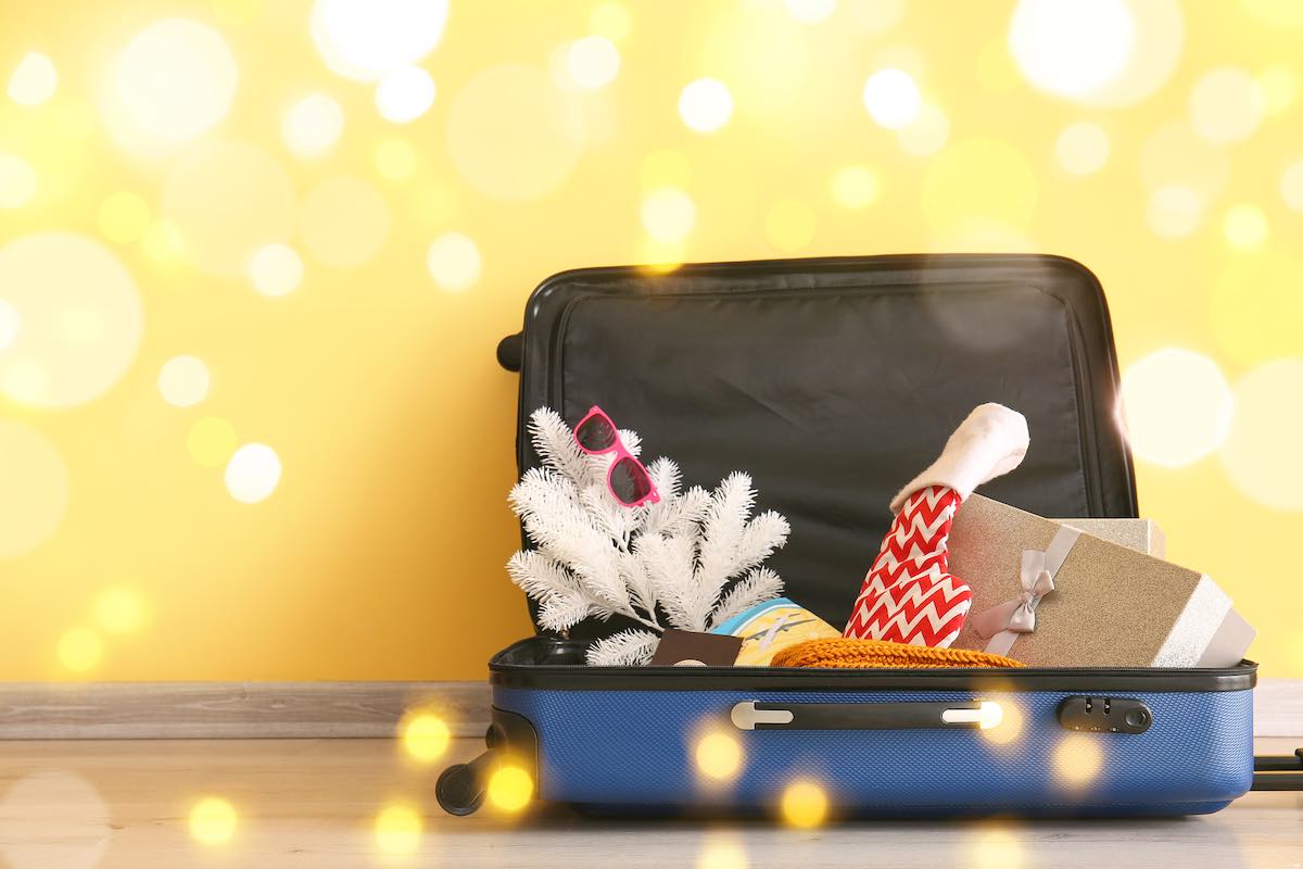 Find out what to pack for Hawaii in December recommended by top Hawaii blog Hawaii Travel Spot! Image of Suitcase with fir tree, Santa sock, sunglasses and gift box near yellow wall. Christmas vacation concept