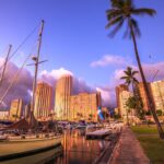 Find out the best Oahu sunset cruises worth booking according to top Hawaii blog Hawaii Travel Spot! Image o Beautiful panorama of sailing boats docked at the Ala Wai Harbor the largest yacht harbor of Hawaii and Honolulu skyline at twilight. On background, a luxurious hotel near Waikiki beach in Honolulu.
