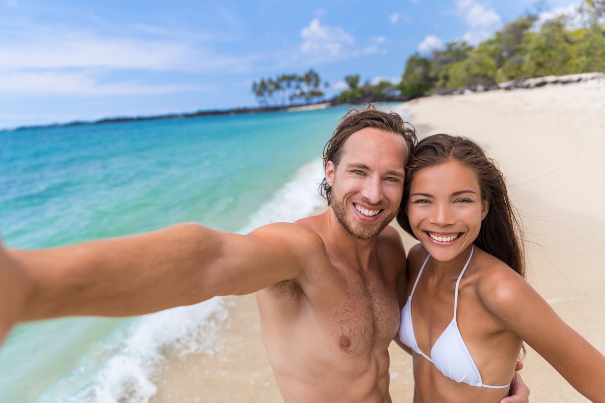 Find out the best Hawaii honeymoon quotes for Instagram compiled by top Hawaii blog Hawaii Travel Spot! Image of Happy seflie couple taking picture with smartphone on beach vacation smiling at camera. Asian woman, Caucasian man. Young multiracial people having fun together in summer travel destination.