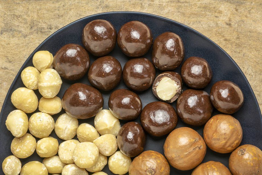 Image of macadamia nuts in shells, shelled and dipped in dark chocolate on a black plate