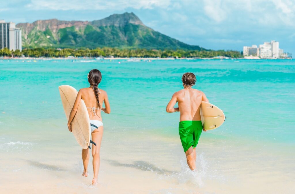 Oahu Travel Guide and 7-Day Oahu Itinerary by top Hawaii blog Hawaii Travel Spot. Image of Hawaii Honolulu couple surfers going surfing on waikiki beach with surfboards running in water. Healthy active sport lifestyle fitness people at diamond head mountain landscape.